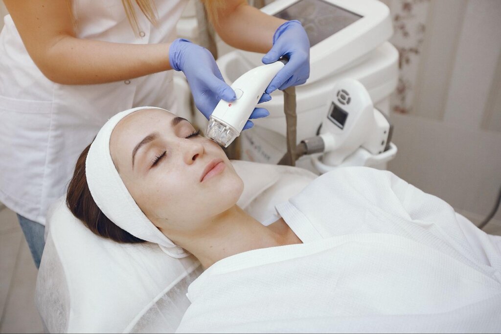 Žena leží v salóne pod prístrojom, ktorý jej ošetruje jazvy po akné na tvári